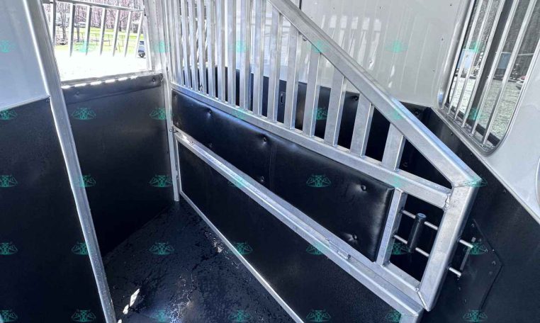 Interior of a horse trailer showing a metal divider with vertical bars and a padded wall along the stall, forming a narrow aisle with a wet, rubberized floor.