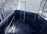 Interior of a white horse trailer with black padded walls and metal dividers along one side; a narrow doorway at the far end, bar-covered windows, and a wet black rubber floor inside.