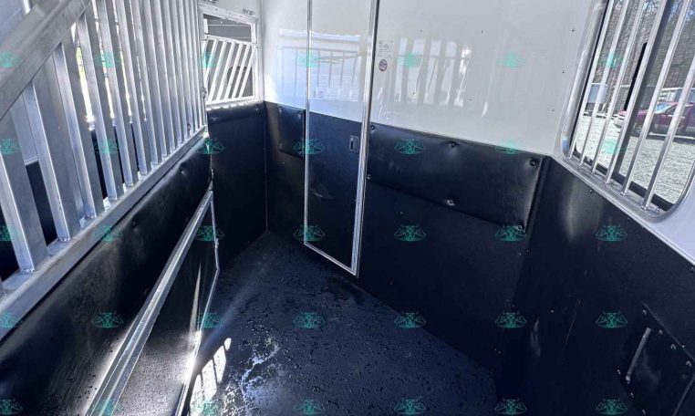 Interior of a white horse trailer with black padded walls and metal dividers along one side; a narrow doorway at the far end, bar-covered windows, and a wet black rubber floor inside.