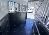 Interior of a horse trailer with a padded black wall on the left, metal divider on the right, barred windows, and a blue non-slip floor; door open to outdoor trailers.