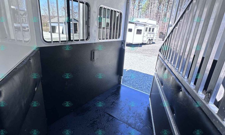 Interior of a horse trailer with a padded black wall on the left, metal divider on the right, barred windows, and a blue non-slip floor; door open to outdoor trailers.