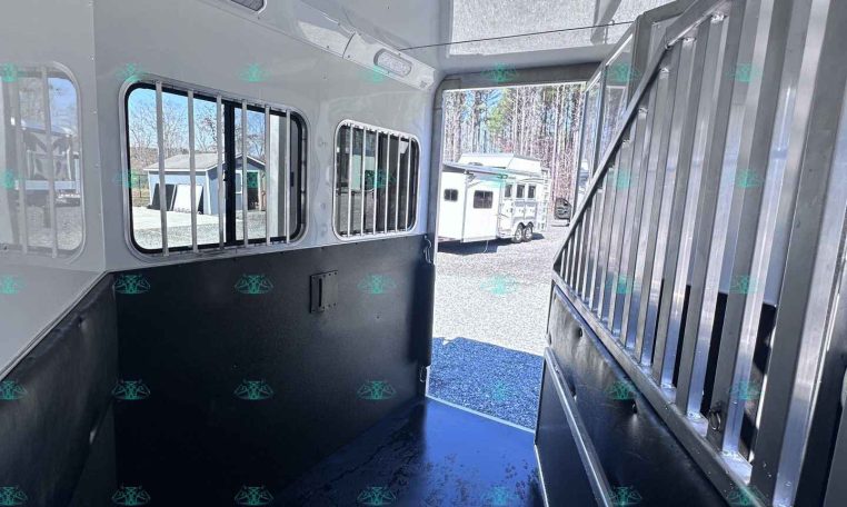 Interior of a white horse trailer with black padded walls and metal stalls, doorway open to bright outside light.