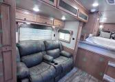 Interior of a RV/trailer living area with a gray leather sofa, wood cabinets above, and a sleeping bunk area in the back.