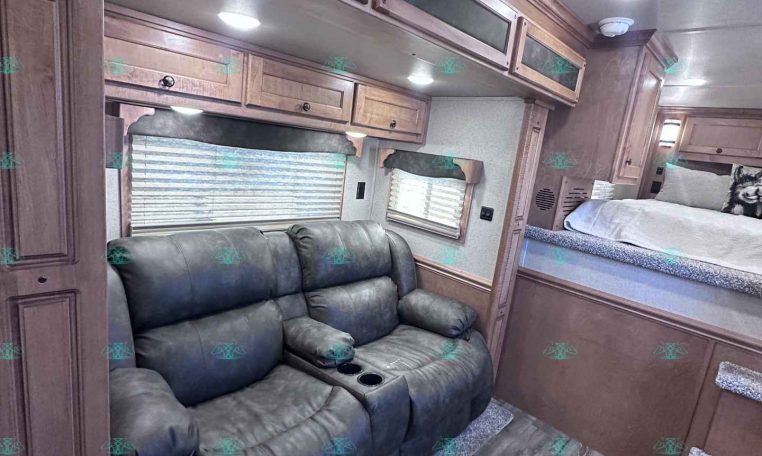 Interior of a RV/trailer living area with a gray leather sofa, wood cabinets above, and a sleeping bunk area in the back.