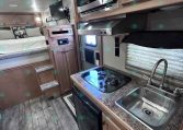 Compact RV kitchen with stainless sink, two-burner stove, and microwave above, plus wooden cabinets and countertop. Bed area visible in background.
