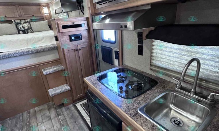 Compact RV kitchen with stainless sink, two-burner stove, and microwave above, plus wooden cabinets and countertop. Bed area visible in background.