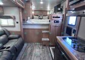 Interior of a modern RV living area: sofa on left, kitchen on right, bed at rear with small steps leading up, ceiling AC unit above.