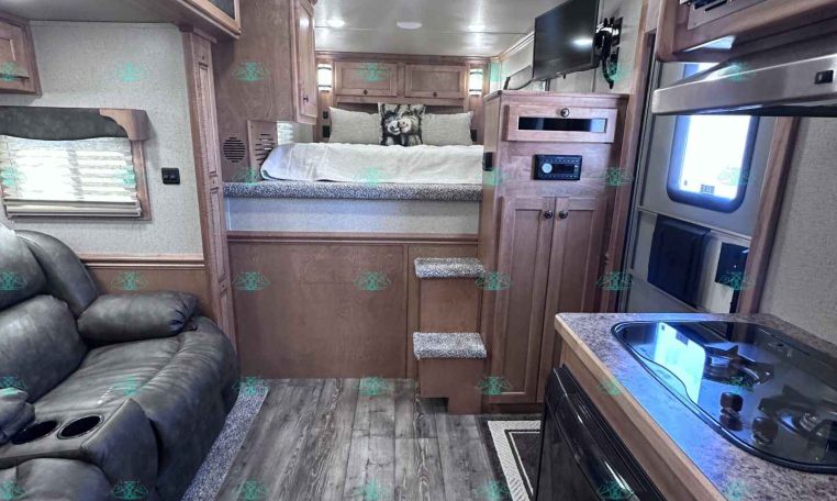Interior of a modern RV living area: sofa on left, kitchen on right, bed at rear with small steps leading up, ceiling AC unit above.