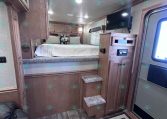 RV bedroom with a raised platform bed, wooden cabinets, and carpeted stairs leading up to the bed