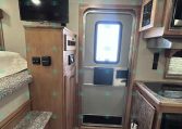 Interior of a compact RV: stairs on the left, a flat-screen TV on a wooden cabinet, and an exterior door with an EXIT label toward the back, with a kitchenette on the right and a fire extinguisher nearby.
