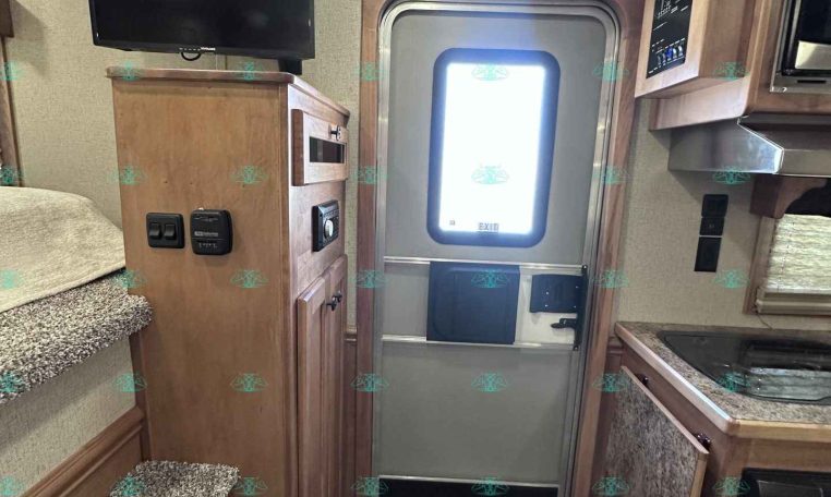 Interior of a compact RV: stairs on the left, a flat-screen TV on a wooden cabinet, and an exterior door with an EXIT label toward the back, with a kitchenette on the right and a fire extinguisher nearby.