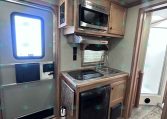 Interior of a compact RV kitchen with microwave above a two-burner stove and sink, and a black mini fridge beside the cabinet.