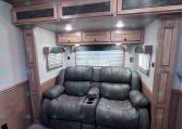 Interior of an RV living area with a dark leather sofa, wood overhead cabinets, and windows with blinds.