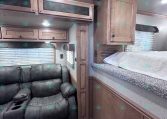 Interior of an RV/travel trailer with brown wood cabinets, a black leather recliner sofa, and a raised bunk area along the right wall.