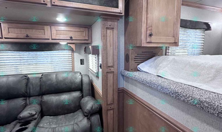 Interior of an RV/travel trailer with brown wood cabinets, a black leather recliner sofa, and a raised bunk area along the right wall.