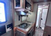 Compact RV kitchen with a two-burner stove, sink, microwave, and wooden cabinets; Merhow branding on the upper cabinet.