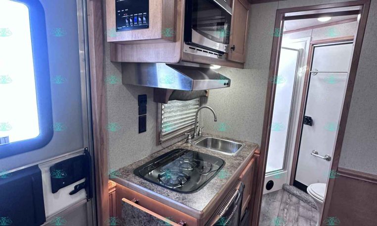 Compact RV kitchen with a two-burner stove, sink, microwave, and wooden cabinets; Merhow branding on the upper cabinet.