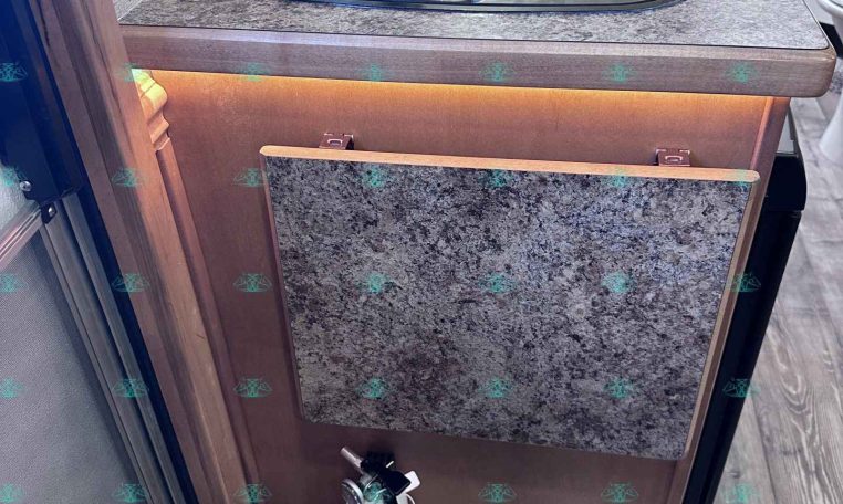 Compact trailer kitchen with granite countertop, sink, and glass-covered stove. Lower cabinet has a fold‑down panel; a fire extinguisher sits on the floor nearby, and a striped rug lies in front.