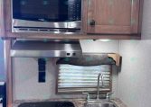 RV kitchen: microwave mounted above a two-burner cooktop, granite counter, sink, and wooden cabinets with a small window and blind.
