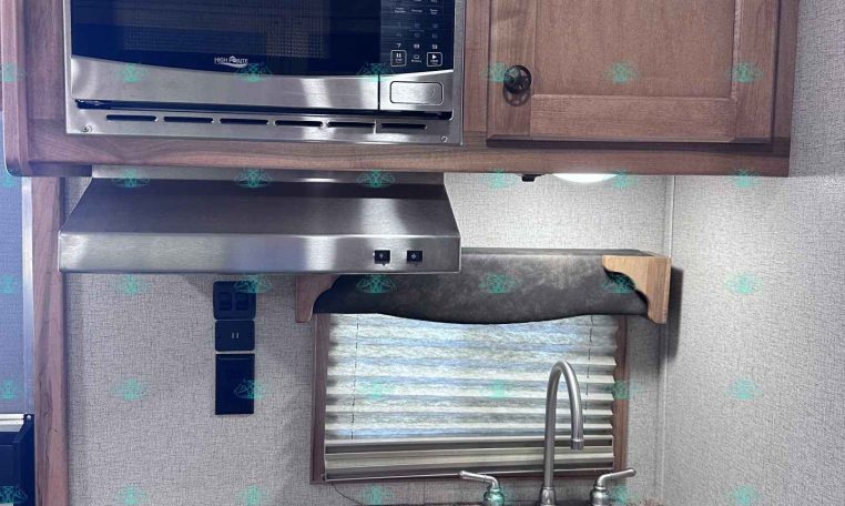 RV kitchen: microwave mounted above a two-burner cooktop, granite counter, sink, and wooden cabinets with a small window and blind.