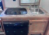 Compact RV kitchen with a two-burner glass cooktop, stainless sink, and a black Dometic dishwasher beneath the counter.