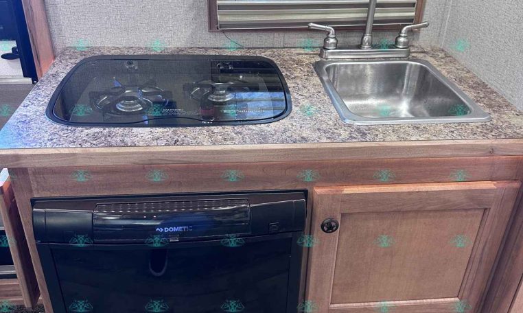 Compact RV kitchen with a two-burner glass cooktop, stainless sink, and a black Dometic dishwasher beneath the counter.