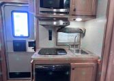 Compact RV kitchen with stainless microwave above a two-burner stove, granite countertop, and small sink under wood cabinetry.