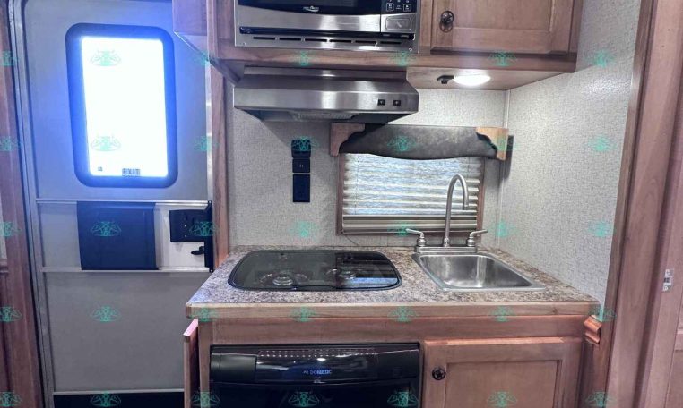Compact RV kitchen with stainless microwave above a two-burner stove, granite countertop, and small sink under wood cabinetry.