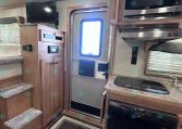 Compact RV interior showing a kitchenette with stove, fridge, and microwave, wood cabinets, and a doorway to the outside on the right.—stair steps on the left and a small TV mounted above the cabinet