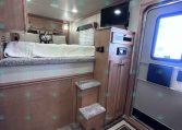 Interior of a travel trailer bedroom with a raised bed, wood cabinets, stairs, and a wall-mounted TV