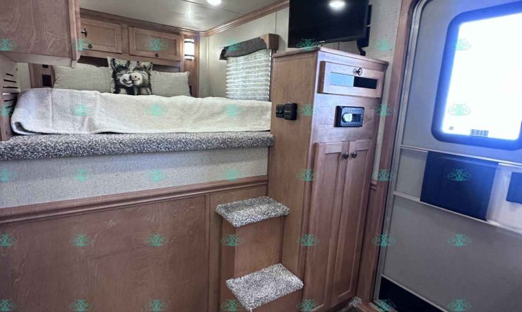 Interior of a travel trailer bedroom with a raised bed, wood cabinets, stairs, and a wall-mounted TV
