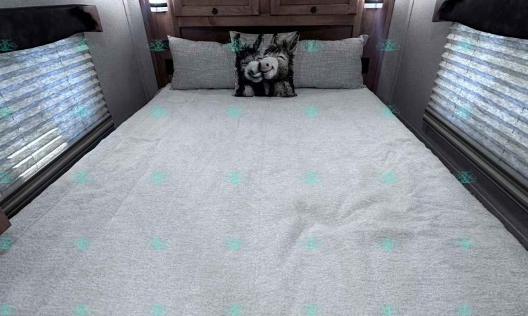 Cozy trailer bedroom with a large bed, gray bedding, and a wooden headboard cabinet against the back wall with two side lamps and windows with blinds on each side