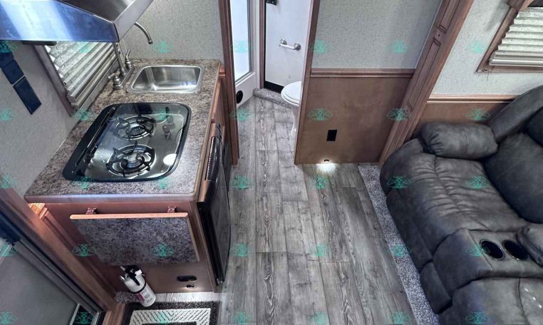 Compact RV kitchen with a two-burner stove, stainless sink, microwave above, and granite-like countertops; doorway to a small bathroom visible in the background.