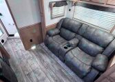RV living area with a plush gray reclining sofa, wood cabinets, and large windows with blinds along the wall.