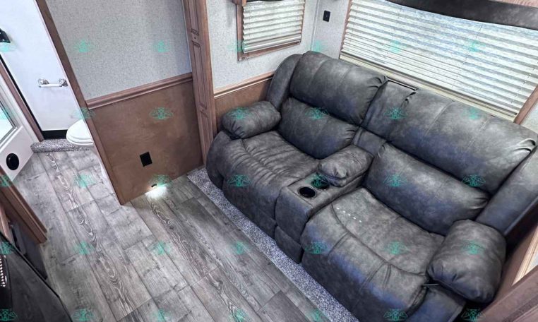 RV living area with a plush gray reclining sofa, wood cabinets, and large windows with blinds along the wall.