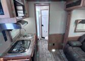 Interior of a compact RV kitchen with a stove, sink, microwave, and overhead cabinets; a sofa is visible to the right and a bathroom door at rear.