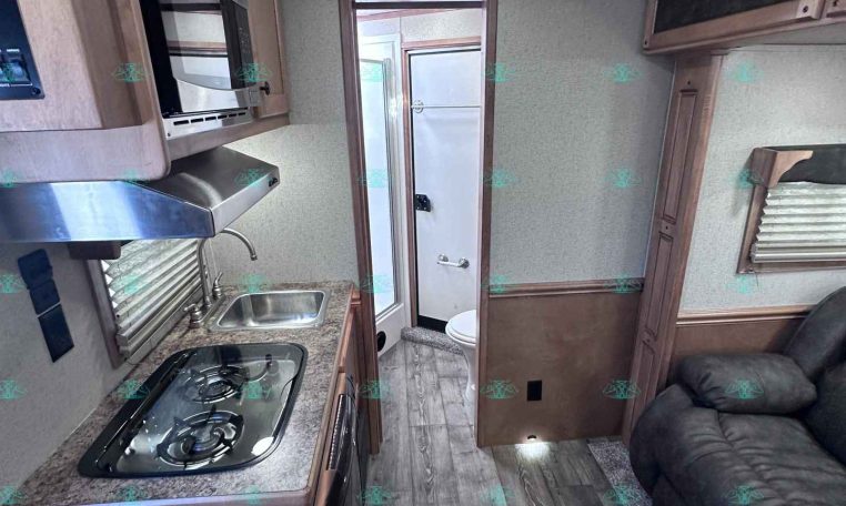 Interior of a compact RV kitchen with a stove, sink, microwave, and overhead cabinets; a sofa is visible to the right and a bathroom door at rear.