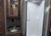 Compact RV bathroom with a wooden vanity, stainless sink, and mirrored medicine cabinet beside a frosted-glass shower stall with a textured door.