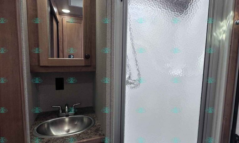 Compact RV bathroom with a wooden vanity, stainless sink, and mirrored medicine cabinet beside a frosted-glass shower stall with a textured door.