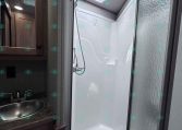 Shower stall in an RV bathroom with a square skylight, handheld showerhead, and frosted glass door beside a wooden vanity and sink countertop.