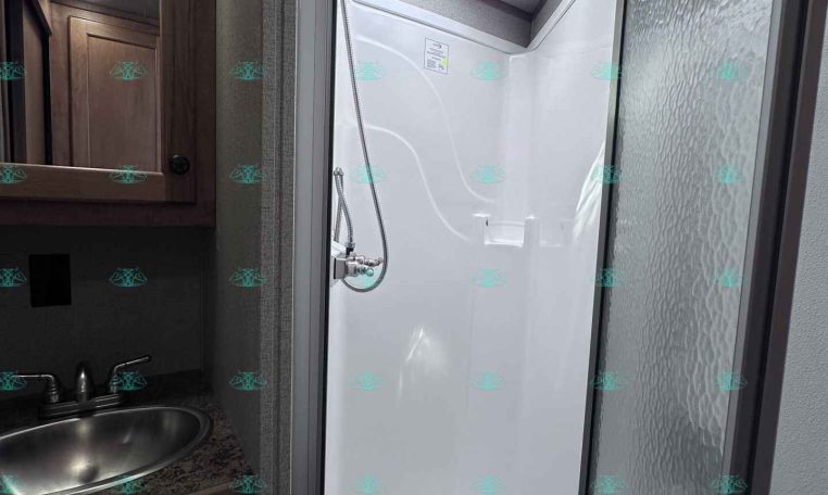 Shower stall in an RV bathroom with a square skylight, handheld showerhead, and frosted glass door beside a wooden vanity and sink countertop.
