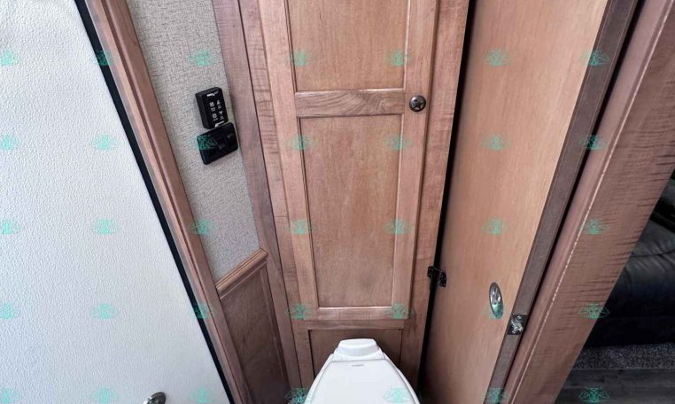 Compact RV bathroom with a white toilet flanked by wood cabinets and a panel door to the right.