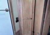 Small RV bathroom with a white toilet and tall wooden cabinet, adjacent panel and door in a wood-tone interior.