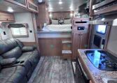 Interior of a modern RV: kitchen counter with stove and sink on the right, leather sofa on the left, and a bedroom loft at the far end.