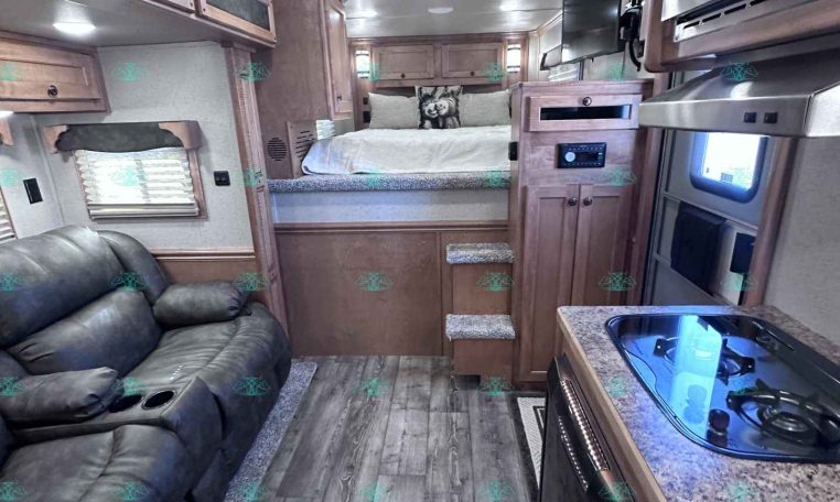 Interior of a modern RV: kitchen counter with stove and sink on the right, leather sofa on the left, and a bedroom loft at the far end.