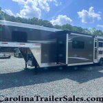 Large gray-and-white fifth-wheel trailer with an extended slide-out parked on a gravel lot, blue sky and trees in the background.