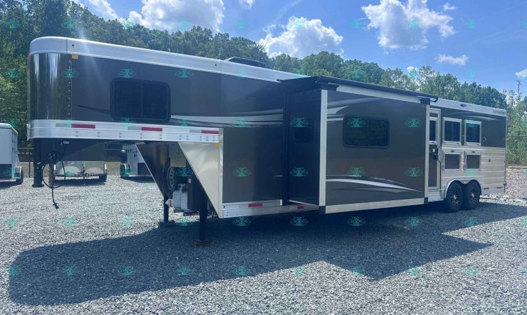 Large gray-and-white fifth-wheel trailer with an extended slide-out parked on a gravel lot, blue sky and trees in the background.