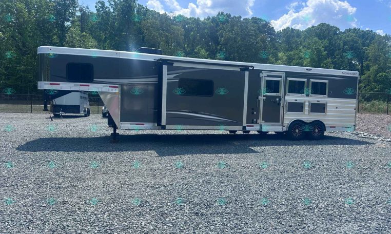 Long dark gray fifth-wheel trailer with a slide-out, parked on a gravel lot with a treeline in the background.