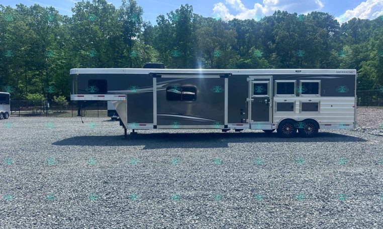 Long gray Merhow horse trailer parked on a gravel lot with green trees and a blue sky in the background.