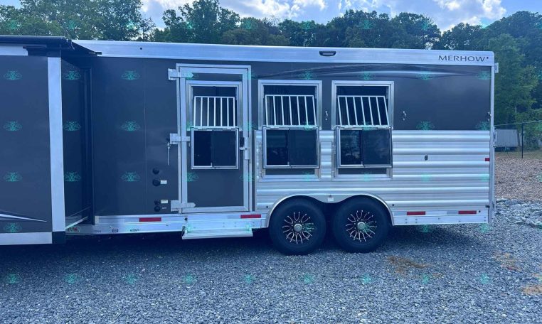 Merhow horse trailer with three axles, dark gray exterior, and side stalls with barred windows, parked on gravel with trees in the background.